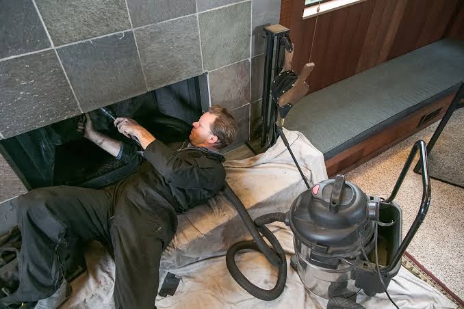 Chimney technician lying on a drop cloth reaching up into the smoke chamber of a slate-tiled fireplace with a vacuum nearby